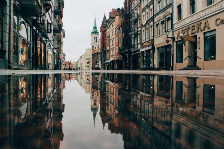 Home A tranquil street in Toruń, Poland, with historic architecture reflecting in a puddle.