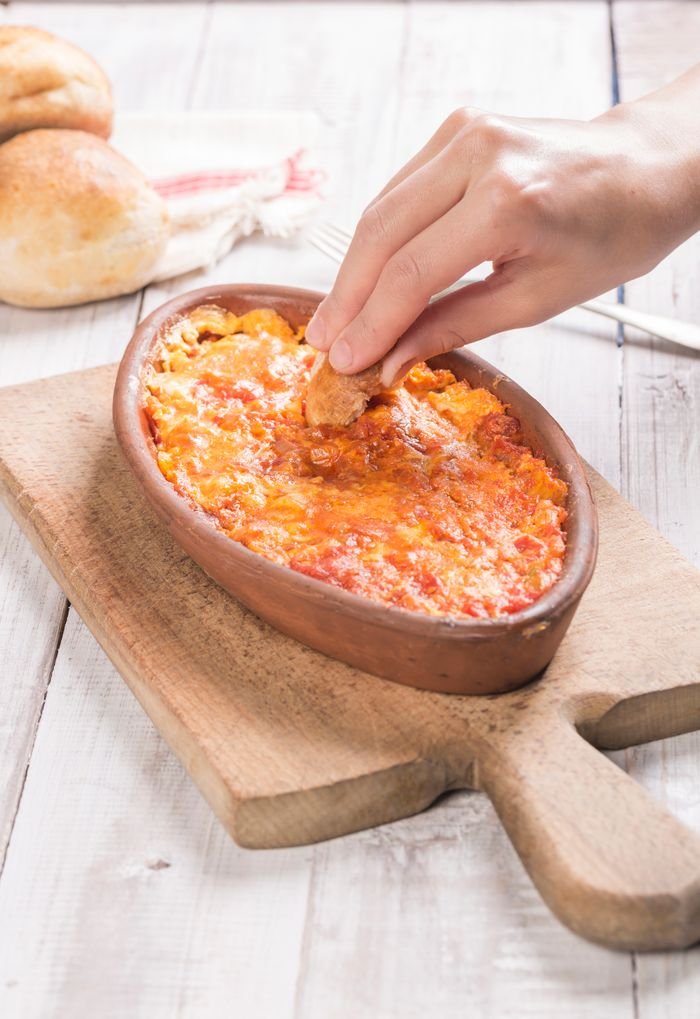 A hand dips bread into a traditional Turkish Menemen dish, served on a wooden board.